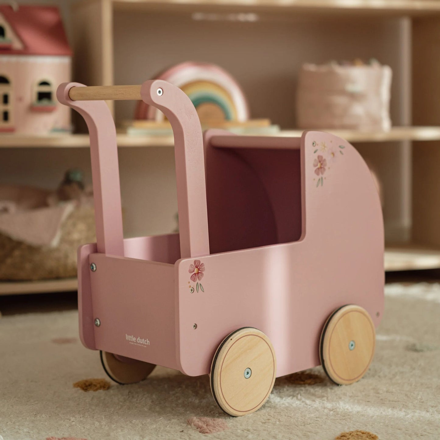 Pink toy pram with wooden wheels and handle in a child's playroom setting.