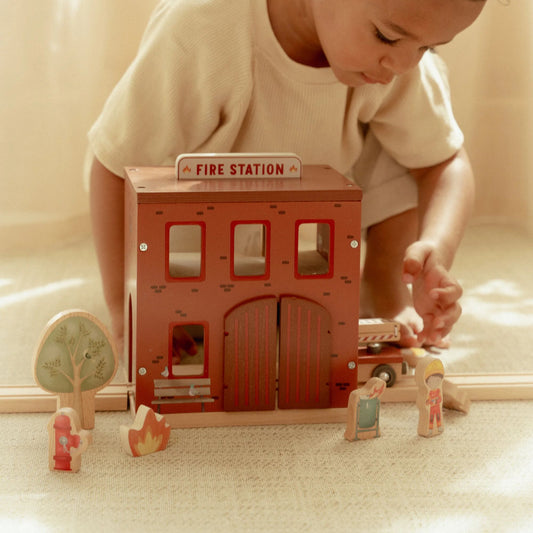 Child playing with a wooden toy fire station on a light-colored floor.