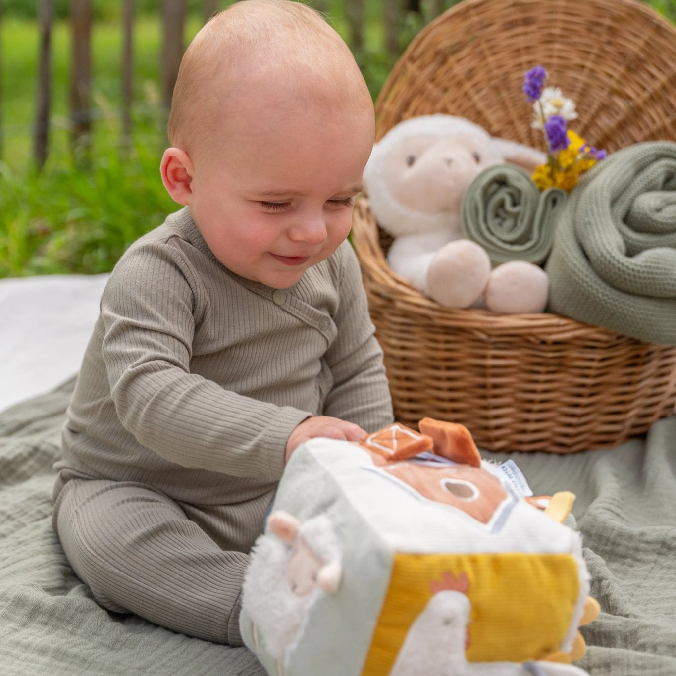 Baby playing with a plush toy outdoors, surrounded by a basket of stuffed animals and blankets.