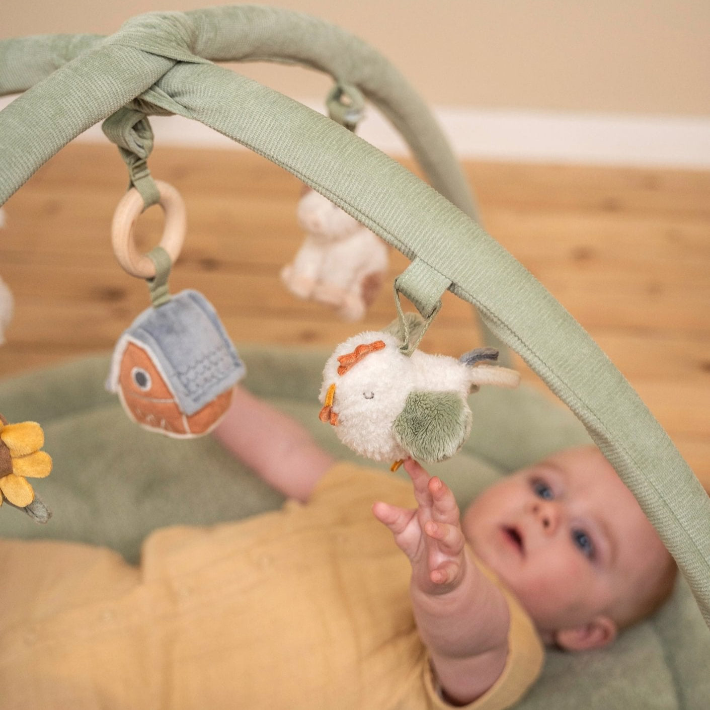 Baby lying on a green play mat with toys, surrounded by soft toys on a wooden floor.