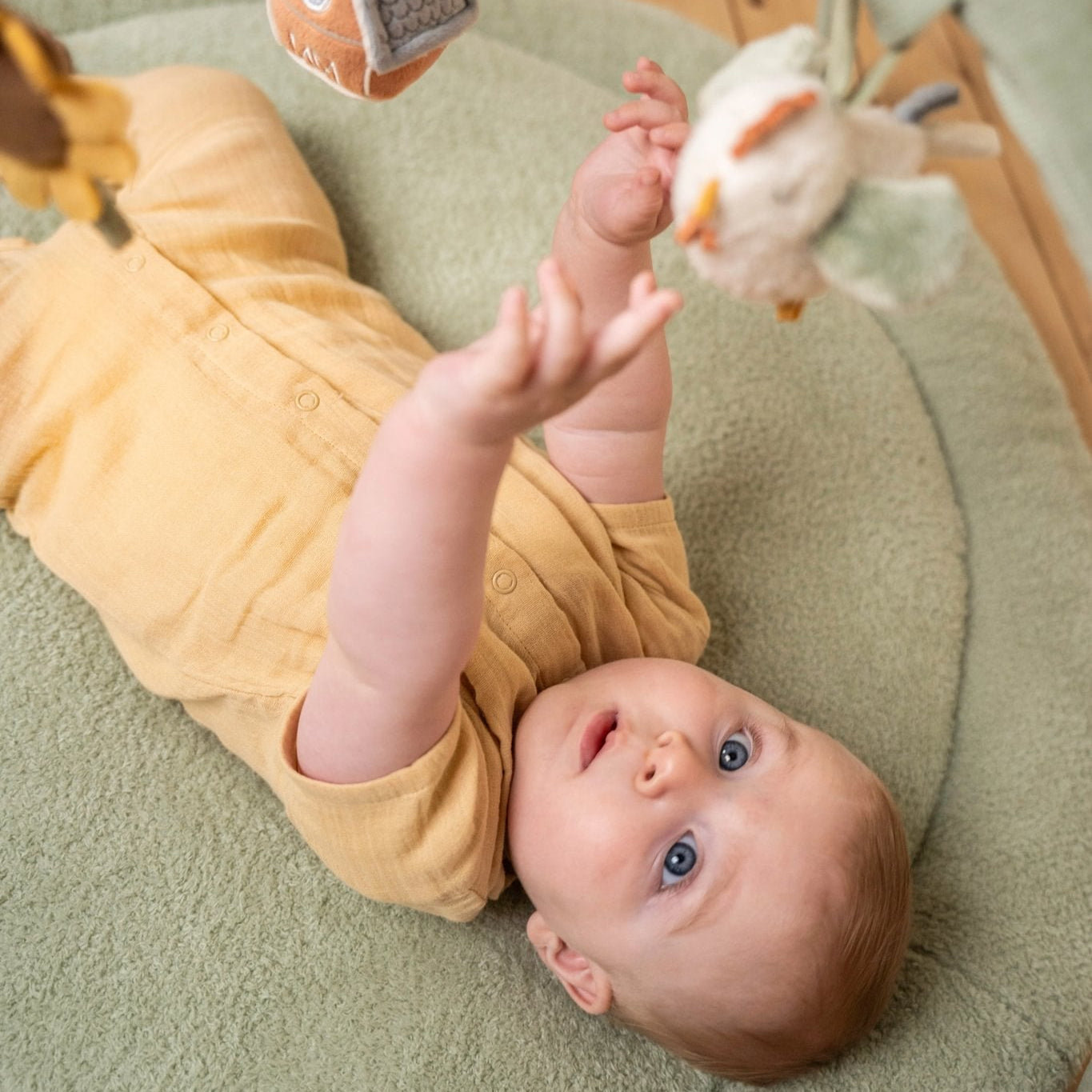 Baby lying on a green cushion with toys around, wearing a yellow outfit.