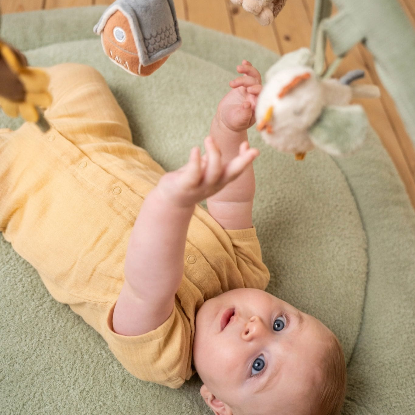 Baby lying on a green cushion with toys around, wearing a yellow outfit.