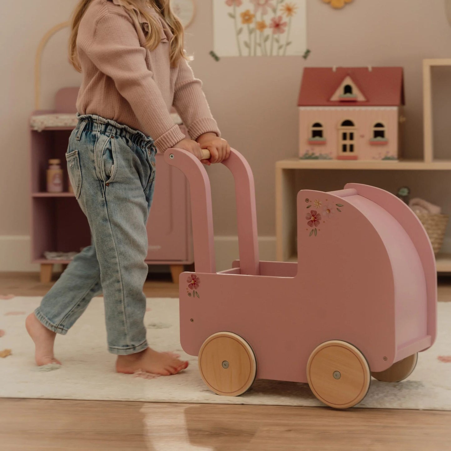 Child pushing a pink doll pram in a room with a toy house in the background