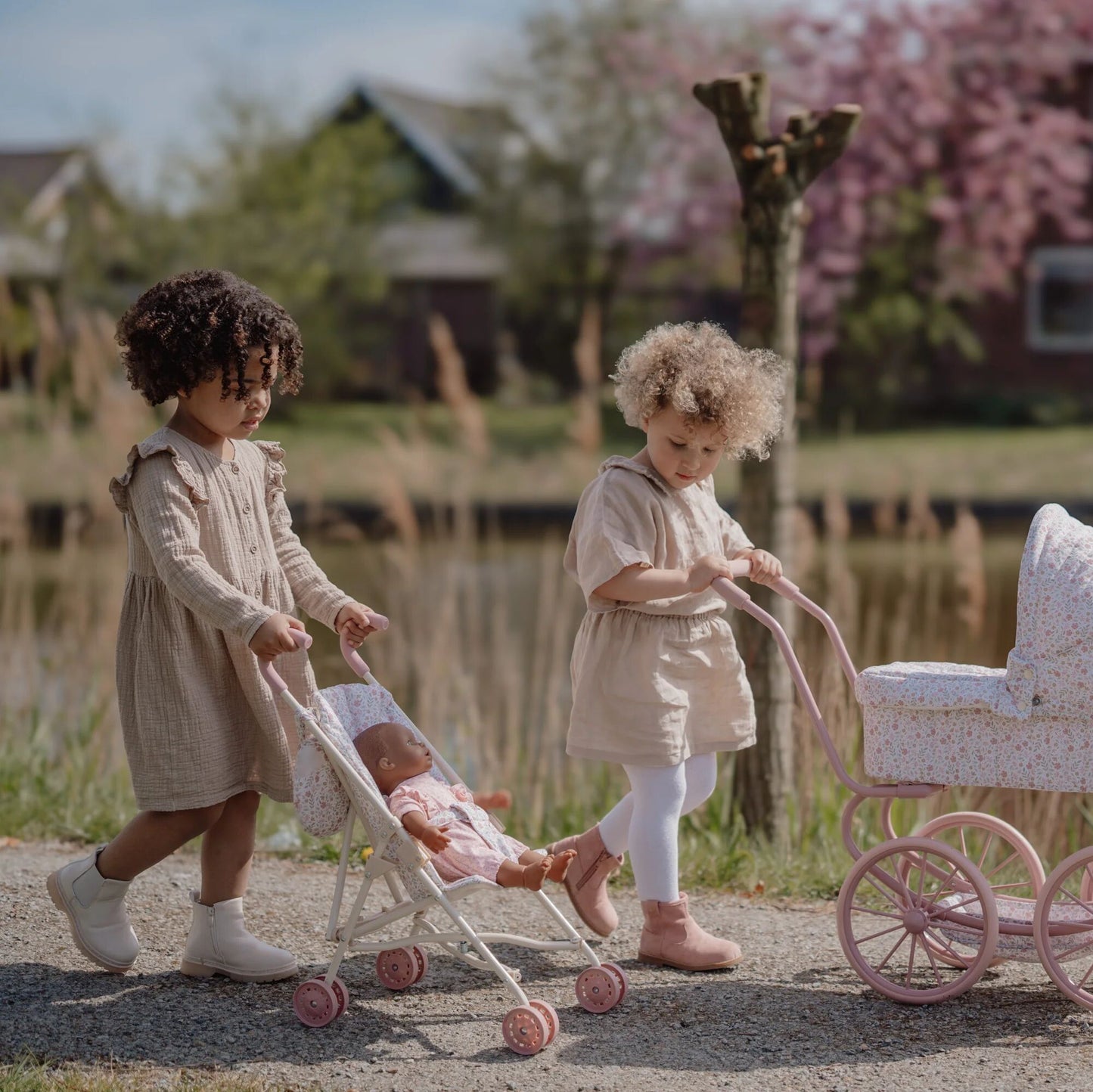 Two young girls playing with dolls and vintage-style strollers in a garden.