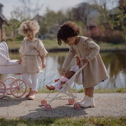 Two young girls playing with dolls and doll strollers by a pond in a park.
